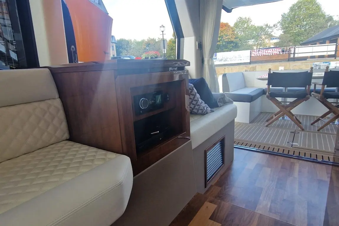 Interior view of a stylish boat lounge with quilted seating, a wooden cabinet with a sound system, and a dining area in the background.